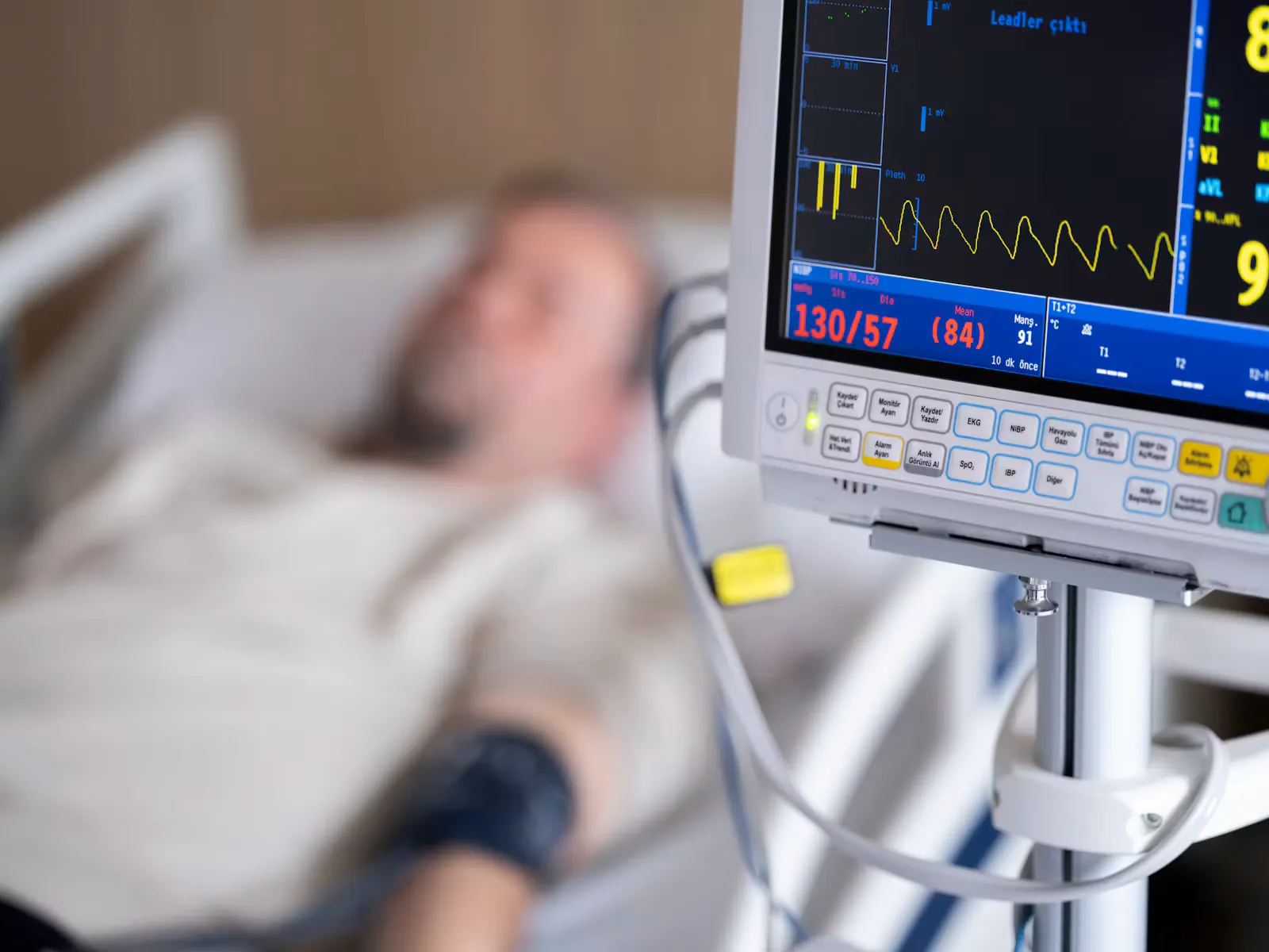A patient monitor showing blood pressure and heart rate next to a hospital bed with a resting patient out of focus